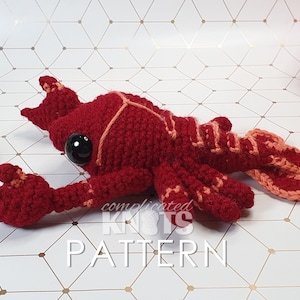 May include: A crocheted lobster amigurumi toy in red and orange. The toy has black eyes and is lying on a white background with a geometric pattern.