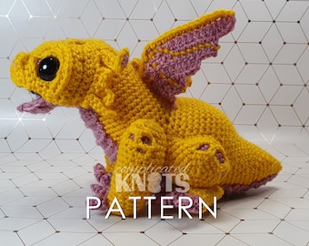 May include: A crocheted yellow dragon with pink wings and a black eye. The dragon is sitting on a white background with a geometric pattern. The text "complicated knots pattern" is visible below the dragon.