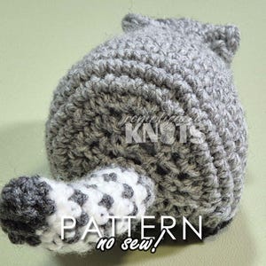 May include: A gray and white crocheted raccoon amigurumi toy with a black nose and white eyes. The toy is made with a no-sew technique. The toy is sitting on a green surface. PATTERN no sew!