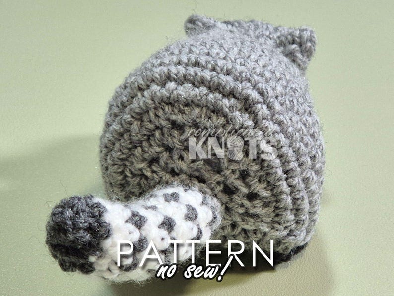 May include: A gray and white crocheted raccoon amigurumi toy with a black nose and white eyes. The toy is made with a no-sew technique. The toy is sitting on a green surface. PATTERN no sew!
