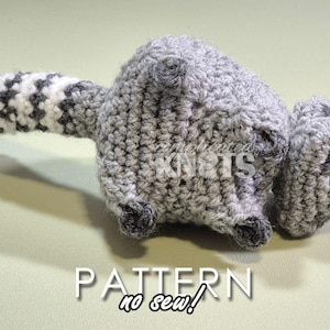 May include: A crocheted raccoon amigurumi toy with a gray and white striped tail. The toy is lying on its side with its head turned to the right. The toy has black eyes and a black nose. The toy is made with a no-sew technique.
