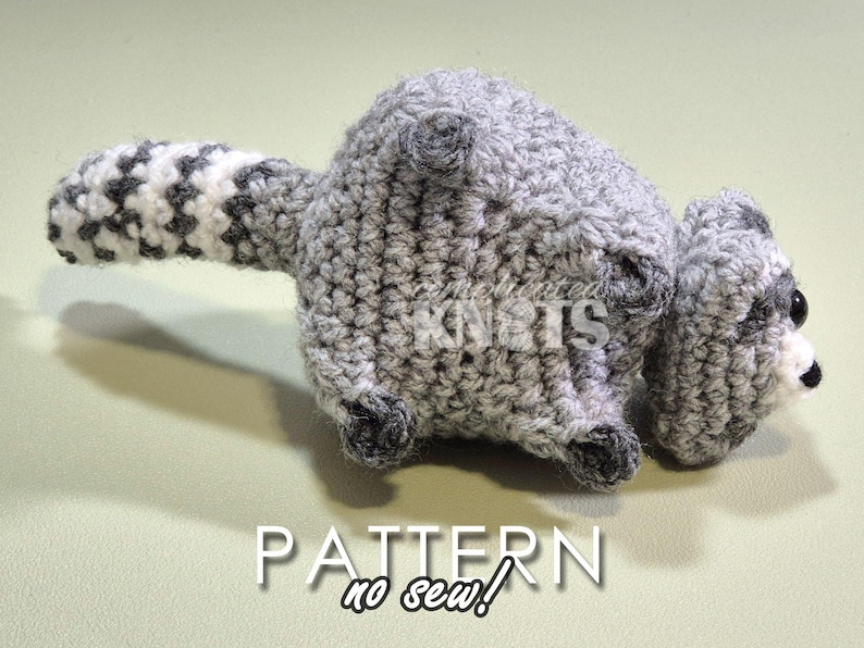 May include: A crocheted raccoon amigurumi toy with a gray and white striped tail. The toy is lying on its side with its head turned to the right. The toy has black eyes and a black nose. The toy is made with a no-sew technique.