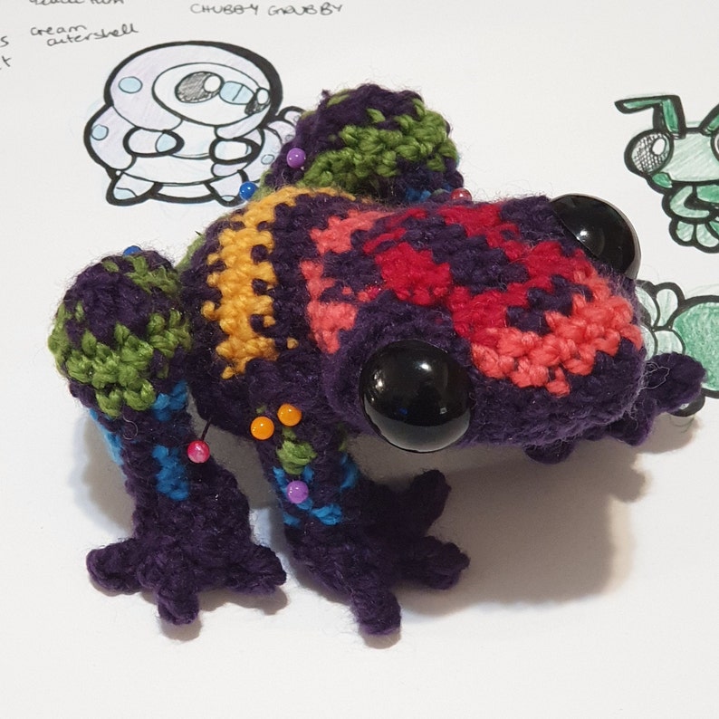 May include: A colorful crocheted frog with large black eyes. The frog is made with different colored yarn, including purple, red, yellow, green, and blue. The frog is sitting on a white surface.