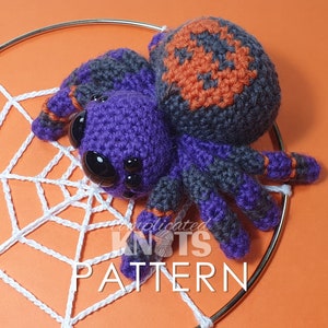 May include: A crocheted purple and grey spider with orange accents sits on a white crocheted web. The spider has large black eyes and is a Halloween-themed craft.