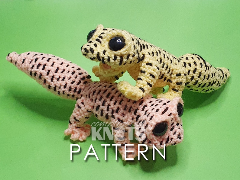 May include: Two crocheted gecko plushies, one yellow with black stripes and one pink with black stripes. The plushies have large black eyes and are posed on a green background.