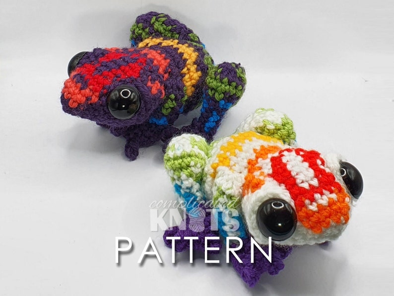 May include: Two colorful crocheted frog amigurumi toys. One frog is purple, red, green, and yellow. The other frog is white, orange, red, and green. Both frogs have black eyes.