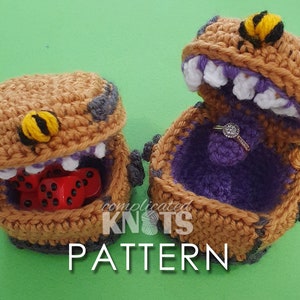 Puede incluir: Dos cajas de ganchillo con un diseño de abeja en la tapa. Una caja está abierta y muestra un interior morado con un anillo dentro. La otra caja está cerrada y muestra dados rojos dentro.