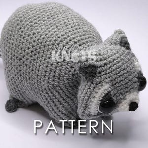 May include: A crocheted raccoon toy in shades of grey, white, and black. The handmade plush animal has a striped tail and button eyes. The word "PATTERN" is displayed in white text below the raccoon.