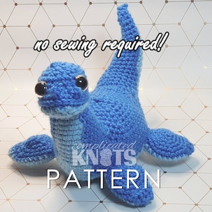 May include: A blue crocheted dinosaur toy with big eyes. The toy is on a white background with a gold geometric pattern. The text "no sewing required!" and "complicated knots pattern" is visible in the image.