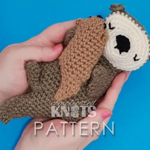 Puede incluir: Un juguete de peluche de nutria tejido a crochet con pelaje marrón y blanco, sosteniendo un pez tejido a crochet marrón en sus patas. El juguete está siendo sostenido en las manos de una persona. El texto "complicated knots pattern" es visible en la imagen.