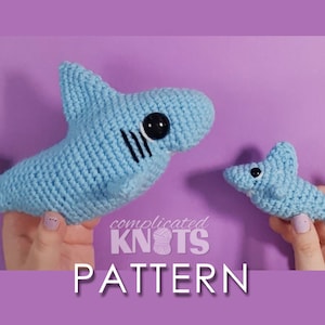 May include: Two crocheted blue shark plushies, one larger than the other, with black eyes and a black stripe pattern. The text "complicated KNOTS PATTERN" is below the plushies.