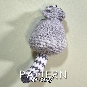 May include: A gray crocheted raccoon with a white and gray striped tail. The raccoon is sitting on a light green surface. The text "PATTERN no sew!" is written below the raccoon.