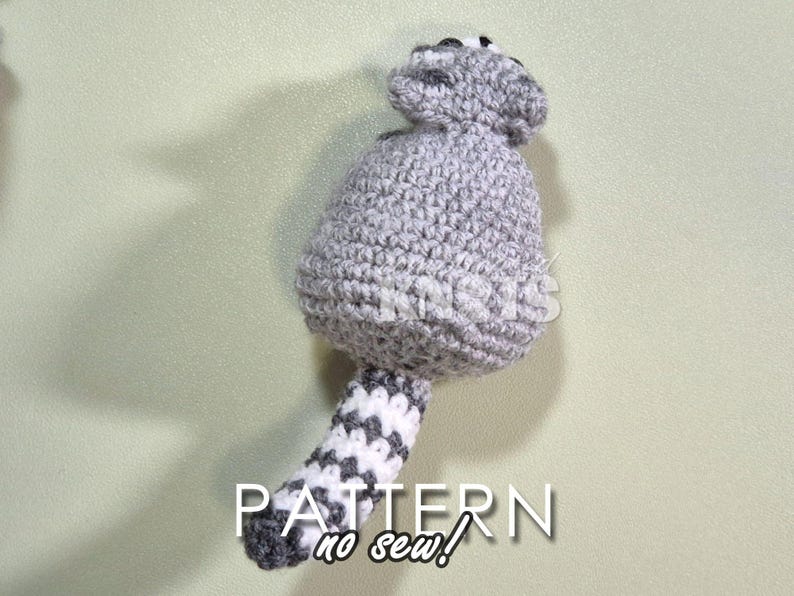 May include: A gray crocheted raccoon with a white and gray striped tail. The raccoon is sitting on a light green surface. The text "PATTERN no sew!" is written below the raccoon.