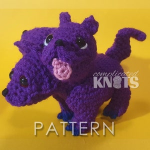 May include: Purple crocheted three-headed dragon amigurumi toy with black eyes and a pink tongue. The toy is sitting on a yellow surface. The text "complicated knots" is visible in the image.