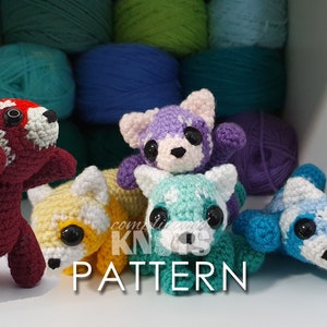 May include: Five crocheted amigurumi animals, including a red panda, a purple cat, a teal cat, a yellow cat, and a blue panda, are arranged on a white surface. The animals are all small and have large eyes. The background is a shelf with balls of yarn in various colors.