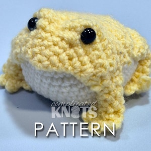 May include: A crocheted yellow frog with black eyes. The frog is sitting on a light blue surface. The text "complicated KNOTS PATTERN" is visible below the frog.