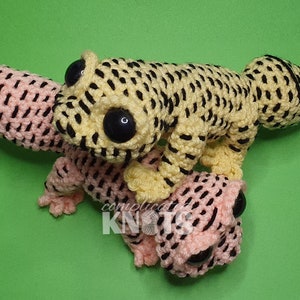 May include: Two crocheted gecko plushies, one yellow with black spots and one pink with black spots. The plushies have large black eyes and are posed on a green background.