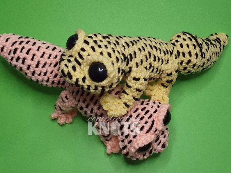May include: Two crocheted gecko plushies, one yellow with black spots and one pink with black spots. The plushies have large black eyes and are posed on a green background.