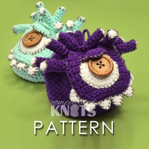 May include: Two round, crocheted monster-like pouches in purple and mint green. Each features a large, central wooden button eye, white crocheted accents, and several small, protruding tentacles on top. The text "complicated KNOTS PATTERN" is visible.