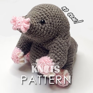 May include: A crocheted mole amigurumi toy with pink accents. The toy is sitting on a white surface. The text "no sew!" is written in black on a white background in the upper right corner of the image. The text "complicated knots pattern" is written in black on a white background in the lower center of the image.