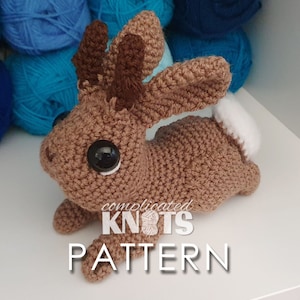 Può includere: Un coniglietto all'uncinetto marrone con corna, occhi grandi e una coda bianca. Il coniglietto è sdraiato su una superficie bianca con gomitoli di lana blu sullo sfondo. Il testo "complicated KNOTS PATTERN" è visibile sotto il coniglietto.