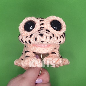 May include: A crocheted pink and black frog with large black eyes. The frog is sitting on a hand.
