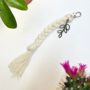 May include: A white braided keychain with a tassel and a small bronze leaf charm. The keychain is attached to a silver key ring.