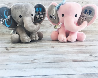 Puede incluir: Dos peluches de elefante gris y rosa con orejas bordadas. El elefante gris tiene el nombre "Emmett" y detalles de nacimiento en una oreja, y el elefante rosa tiene el nombre "Abby" y detalles de nacimiento en la otra oreja.