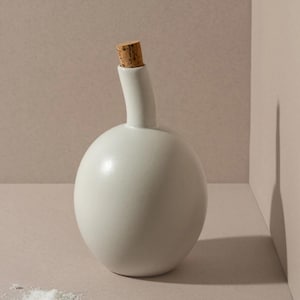 May include: A white ceramic bottle with a cork stopper. The bottle has a rounded base and a slender, curved neck. A small pile of white salt sits near the bottle on a neutral-toned surface.