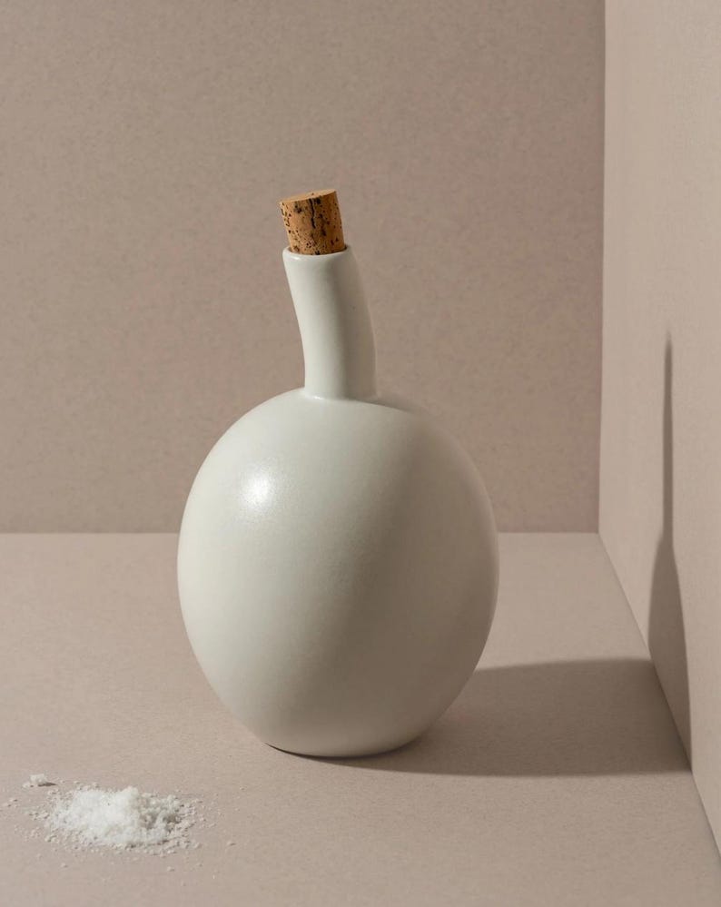 May include: A white ceramic bottle with a cork stopper. The bottle has a rounded base and a slender, curved neck. A small pile of white salt sits near the bottle on a neutral-toned surface.