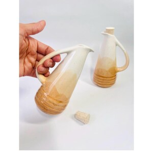 May include: Two ceramic pitchers with a cream and tan color gradient. One pitcher is held by a hand. The pitchers have a handle and a spout. A cork stopper is next to the pitcher. The bottom of the pitchers have a textured design.
