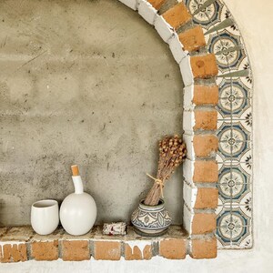 May include: A decorative alcove with a brick and tile archway. A white ceramic cup and bottle with a cork stopper sit on a brick shelf, along with a small patterned vase holding dried flowers. The alcove has a concrete background.