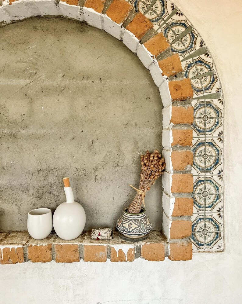 May include: A decorative alcove with a brick and tile archway. A white ceramic cup and bottle with a cork stopper sit on a brick shelf, along with a small patterned vase holding dried flowers. The alcove has a concrete background.
