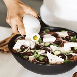 May include: A black bowl filled with a Greek salad, including tomatoes, cucumbers, red onions, olives, and feta cheese. Olive oil is being poured from a white bottle onto the salad, enhancing the fresh ingredients.