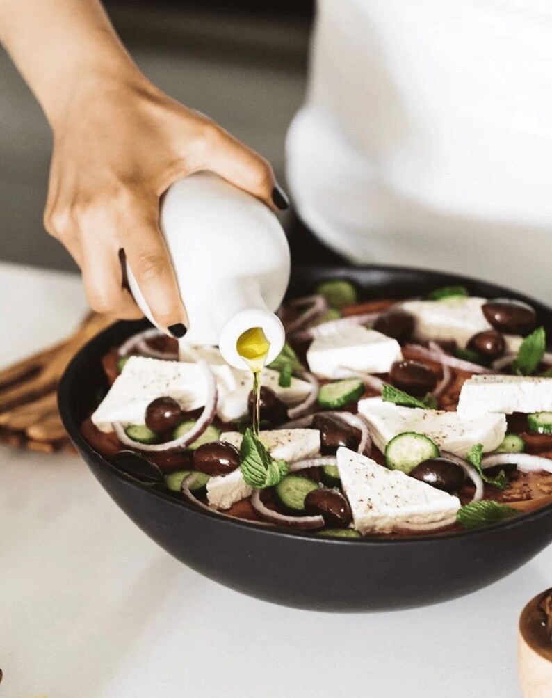 May include: A black bowl filled with a Greek salad, including tomatoes, cucumbers, red onions, olives, and feta cheese. Olive oil is being poured from a white bottle onto the salad, enhancing the fresh ingredients.