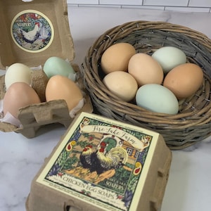 May include: A selection of chicken egg soaps in a range of colours, presented in a cardboard egg box and a wicker basket. The soaps are in shades of beige, light green, and peach. The packaging features the text "Fire Lake Farm" and "Chicken Egg Soaps".