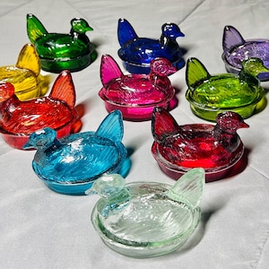 Puede incluir: Una colección de joyeros de cristal con forma de gallina de colores. Las gallinas son de varios colores, como amarillo, verde, azul, morado, rosa y rojo. Cada gallina tiene un diseño detallado y está sentada en un plato pequeño. Estos artículos decorativos son ideales para guardar objetos pequeños.