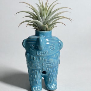 Puede incluir: Una maceta de cerámica turquesa con forma de figura estilizada, con rostro y brazos. La maceta contiene una planta aérea verde con hojas largas y delgadas. La figura tiene detalles grabados y un agujero en la parte delantera.