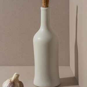 May include: A white ceramic bottle with a cork stopper stands next to a bulb of garlic. The bottle has a slender neck and a rounded base. The garlic bulb is off-white with purple streaks.