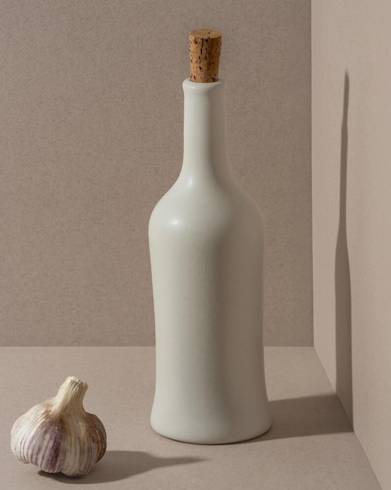May include: A white ceramic bottle with a cork stopper stands next to a bulb of garlic. The bottle has a slender neck and a rounded base. The garlic bulb is off-white with purple streaks.