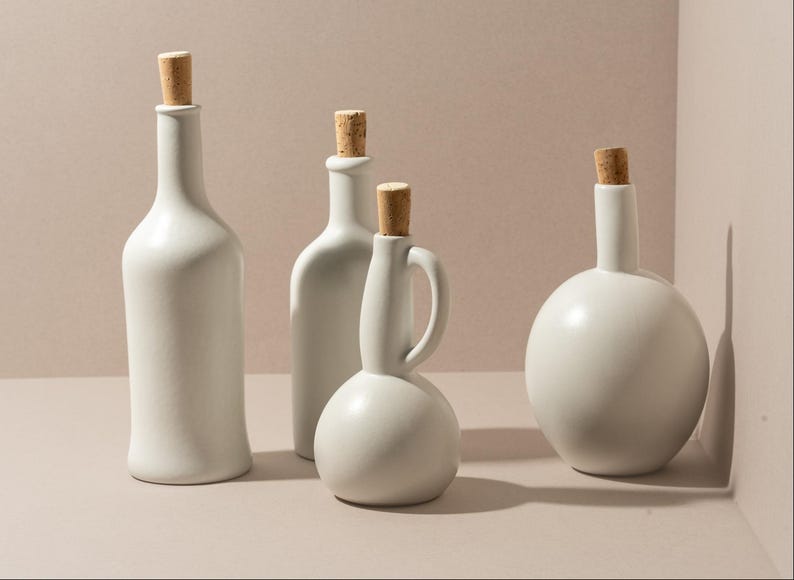 May include: Four white ceramic bottles with cork stoppers. The bottles vary in shape, including a tall, slender bottle, a rounded bottle, and a bottle with a handle. The bottles are arranged on a neutral surface.