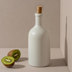 May include: A white ceramic bottle with a cork stopper, next to two halves of a kiwi fruit. The bottle has a smooth, rounded shape. The kiwi fruit is green with black seeds and a brown skin.