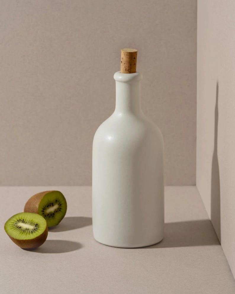 May include: A white ceramic bottle with a cork stopper, next to two halves of a kiwi fruit. The bottle has a smooth, rounded shape. The kiwi fruit is green with black seeds and a brown skin.