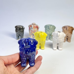 May include: A collection of ceramic figurines in various colors, including blue, yellow, white, and brown. The figurines have a stylized, anthropomorphic design with rectangular features and small legs. The foreground features a blue figurine held in a hand.