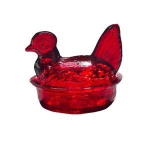 Puede incluir: Un recipiente decorativo de vidrio rojo en forma de gallina sobre un nido. La gallina tiene una cabeza detallada y una cola prominente. El recipiente es probablemente un bombonera o un joyero.