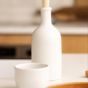 May include: A white ceramic bottle with a cork stopper, a small white bowl, and a portion of a brown dish. The bottle is tall and slender, and the bowl is rounded. The items are on a white surface.