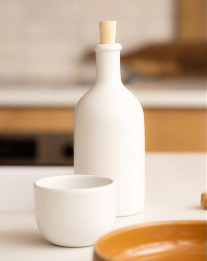 May include: A white ceramic bottle with a cork stopper, a small white bowl, and a portion of a brown dish. The bottle is tall and slender, and the bowl is rounded. The items are on a white surface.