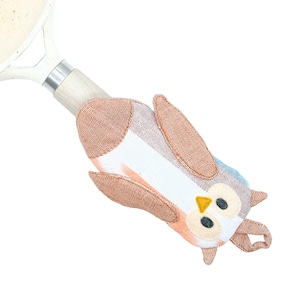 May include: An owl-shaped pot handle cover in shades of brown, white, and blue. The cover is made of fabric and has a loop for hanging. The owl has large black eyes and a yellow beak. The cover is designed to protect hands from heat.