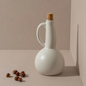 May include: A white ceramic bottle with a handle and a cork stopper. The bottle has a rounded base and a narrow neck. Several brown hazelnuts are scattered near the bottle. The background is a neutral, light-colored surface.