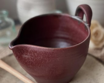 May include: A burgundy-colored ceramic gravy boat with a handle and a pouring spout. The handmade bowl has a textured surface and sits on a neutral-toned plate. A wooden spoon is partially visible in the foreground.